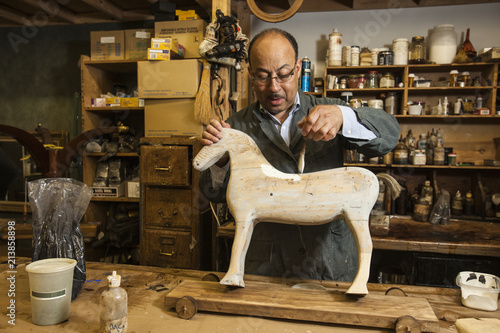 Business owner of antique restoration and wood carving studio at his workbench painting model of wooden horse in New York City, USA photo