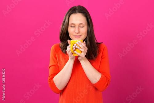Beautiful brunette woman with a yellow cup in hands on a pink background. Drink hot drinks and be healthy.