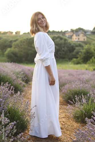 smiling attractive woman in white dress standing in violet lavender field and looking at camera