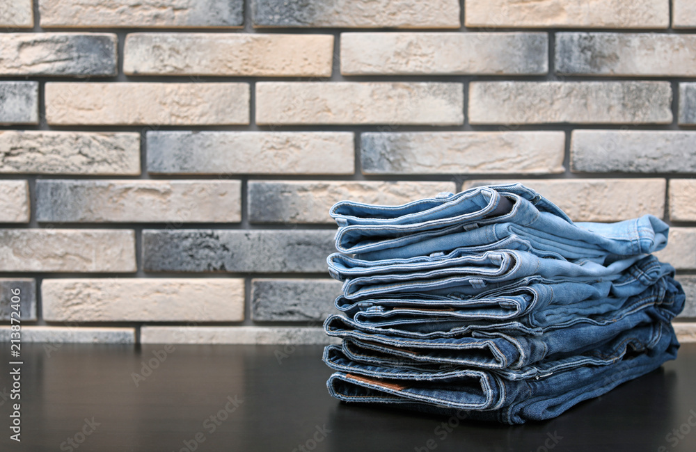 Stack of stylish jeans on table against brick wall