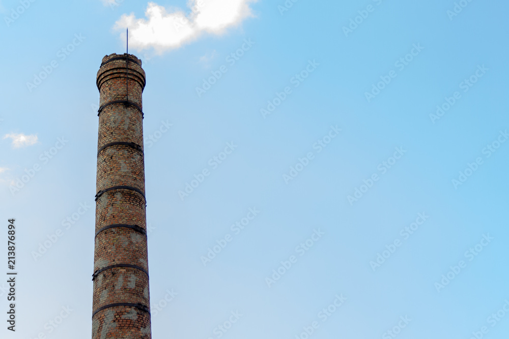 An old dilapidated brick chimney of the plant against the blue sky and clouds. Concept on the theme of ecology and environmental protection from harmful emissions into the atmosphere.