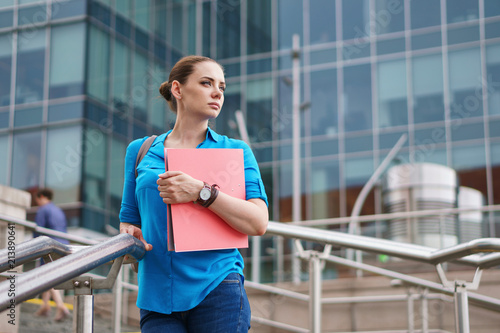 Young attractive girl is holding a folder for documents. The manager stands on the steps of the business center and looks thoughtfully sideways.