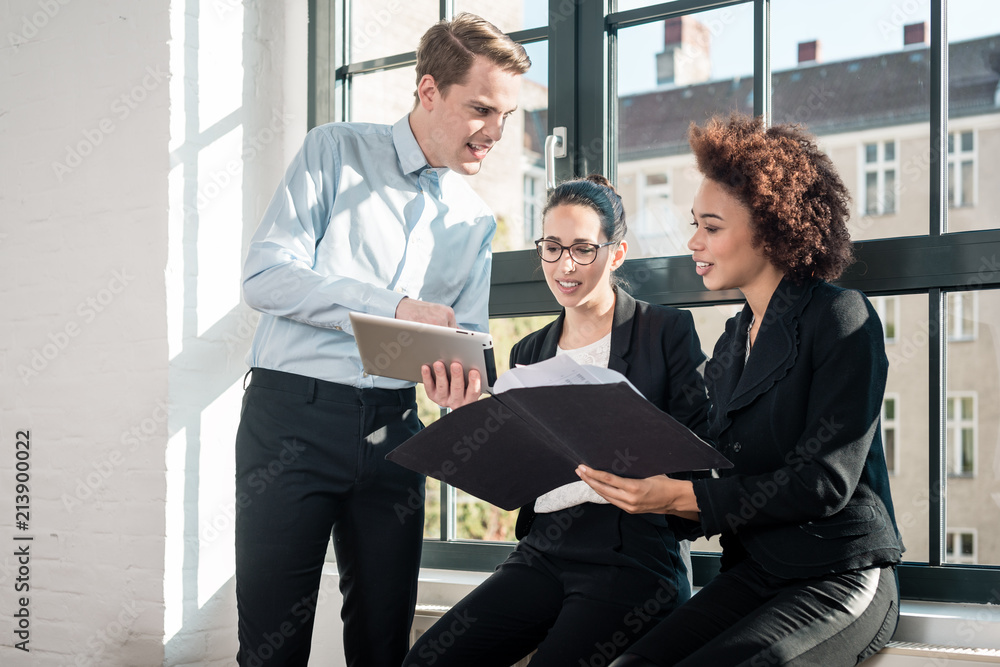 Three young cheerful employees smiling while holding a tablet PC and a folder during break in a modern office Three young cheerful employees smiling while holding a tablet PC and a folder during break in a modern office