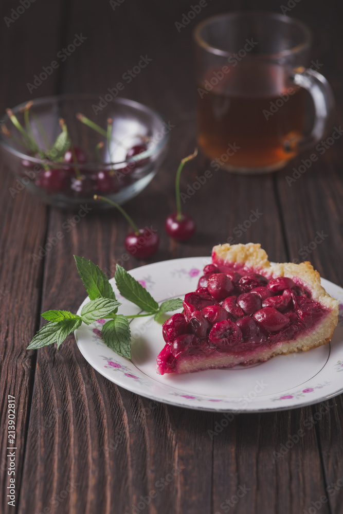 A piece of cherry pie and a sprig of mint lies on a white plate. Behind lie cherry berries and there is a transparent glass cup with tea. All the items lie on an old wooden table. A piece of cherry pie and a sprig of mint lies on a white plate. Behind lie cherry berries and there is a transparent glass cup with tea. All the items lie on an old wooden table.