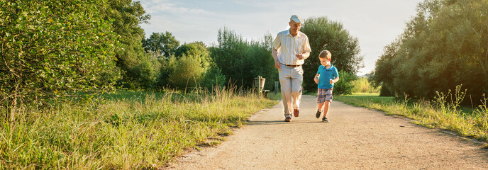 Front view of senior man with hat and happy child running on a nature path. Two different generations concept.