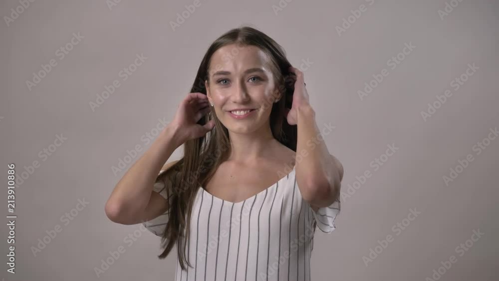 Young charming girl is watching at camera, smiling, touching hair, grey background Young charming girl is watching at camera, smiling, touching hair, grey background