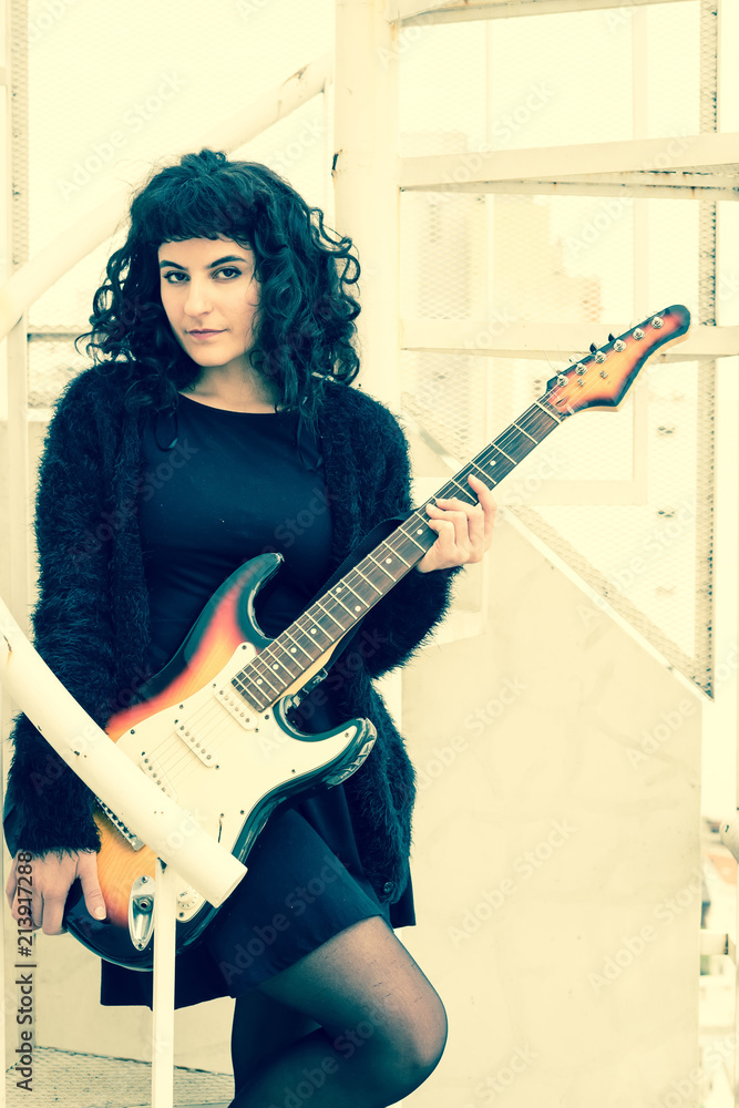 A beautiful young smiling woman in an elegant dress playing on an electric guitar while standing on a metal stairs. A beautiful young smiling woman in an elegant dress playing on an electric guitar while standing on a metal stairs.