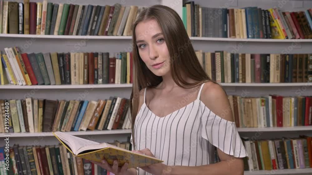 Young attractive girl is reading book in library, watching at camera, smiling Young attractive girl is reading book in library, watching at camera, smiling