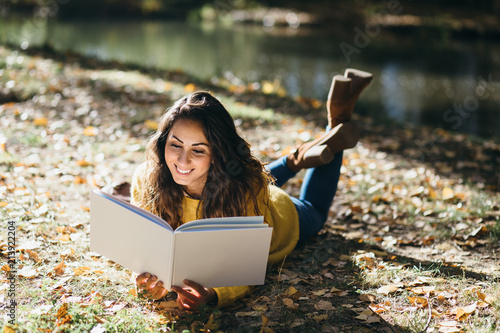 Happy casual woman reading a book at river shore in autumn.
