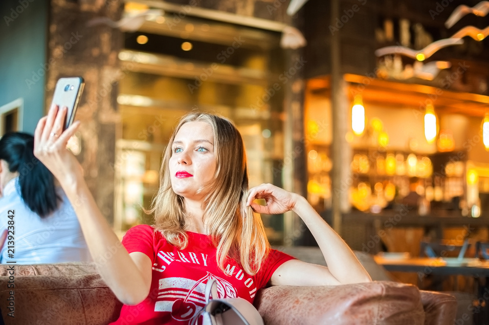 A woman in a cafe at a table does selfie on the phone. A visitor to the restaurant takes pictures of himself on the phone. Woman in red T-shirt and red lipstick