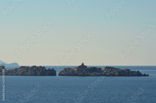 A small light house with a red roof is on an island of rocks. The sky is blue with no clouds.