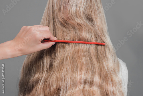 Young woman with comb brushing her wet, blonde, perfect hair on the gray background. Care about beautiful, healthy and clean hair. Beauty salon concept. Side view.