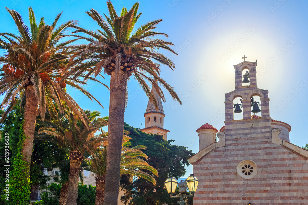Tropical landscape with palms and facade of old historical church and high tower in central district of city Budva, Montenegro, under direct sun