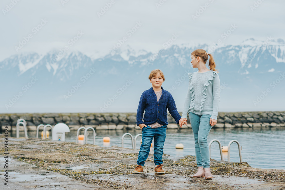 Outdoor portrait of two adorable kids playing together next to lake on a cold day, wearing warm pullover