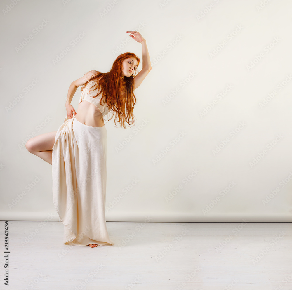 Naklejka premium Dancer posing in studio. Young and beautiful redhead girl in a beige long skirt and top dances and poses in studio. Copy space