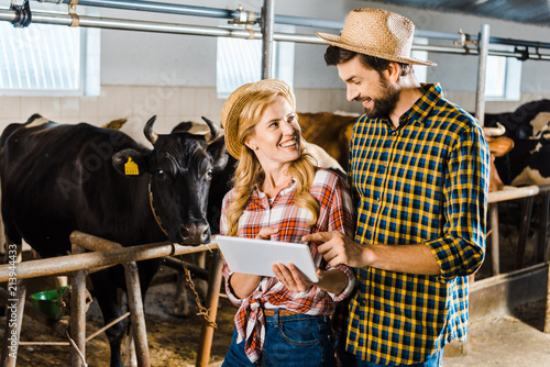 smiling couple of farmers using tablet in stable with cows