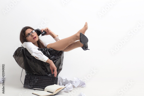 Woman sitting in garbage can uses her shoe as phone. Dorky business lady using her stiletto as phone and pretending to have conversation while sitting in bucket for trash. photo