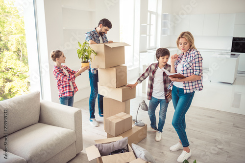 Young happy smiling family four persons unwrapping carton boxes with stuff in light kitchen studio living room, moving to new flat, check list