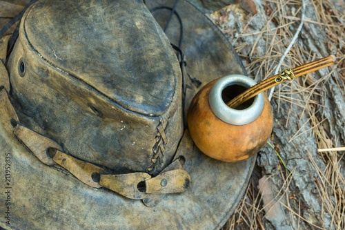 Hand Crafted Artisanal Yerba Mate Tea Leather Calabash Gourd with Straw Hat on Wood Logs in Forest. Travel Wanderlust Concept. Earthy Tones. Traditional Argentinian Latin American Brewing Cup