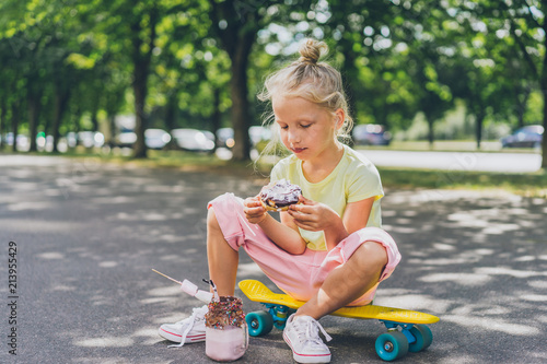 selective focus of little child eating doughnut from dessert while sitting on skateboard at street