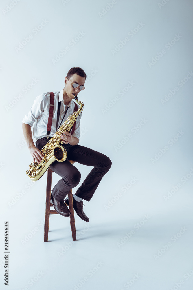 Naklejka premium handsome young jazzman sitting on stool and playing saxophone on grey