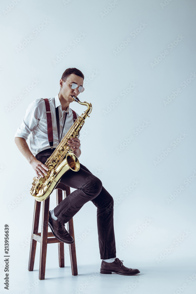 Naklejka premium handsome young performer sitting on stool and playing saxophone on grey