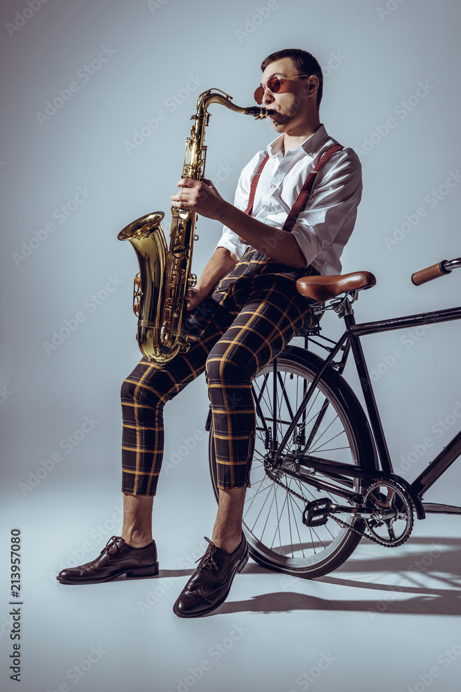 Naklejka premium handsome young performer in sunglasses playing saxophone while sitting on bicycle on grey