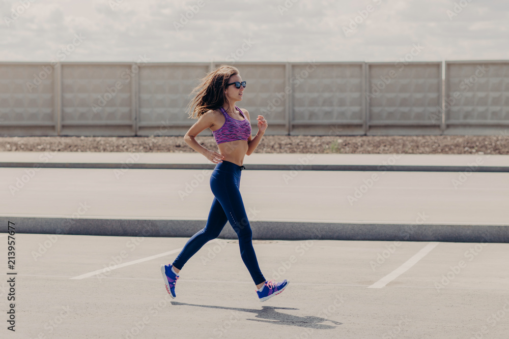 Slim fit female in stylish sportswear warms up during morning run, joggs in open air, being photographed in motion, gets ready for marathon. Active sportswoman has workout before competition