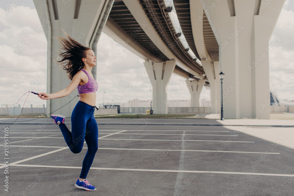 Sideways shot of healthy slim young female with athletic body, jumps with skipping rope, does physical exercises outdoors, wears sportswear. Fitness cardio training with rope. Sport concept. Sideways shot of healthy slim young female with athletic body, jumps with skipping rope, does physical exercises outdoors, wears sportswear. Fitness cardio training with rope. Sport concept.