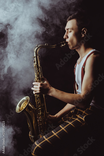 side view of stylish young jazzman sitting and playing saxophone in smoke on black