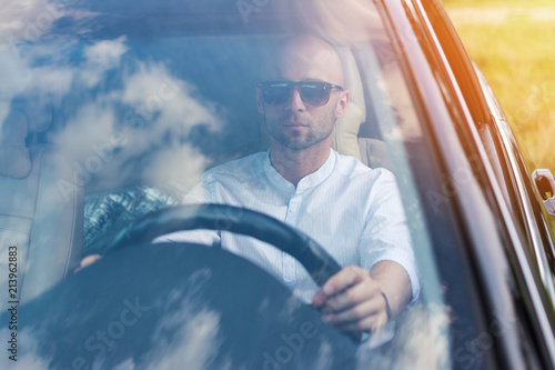 Handsome young man in white shirt and sunglasses driving a car without seat belt. Traffic law violation. Window reflecting sky, natural lighting, no retouch, matte filter.