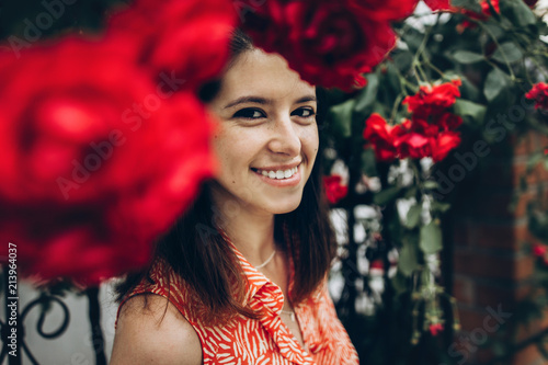 beautiful stylish hipster woman smiling under big red roses in sunny street in summer. beauty of blooming in spring. space for text. atmospheric moment. happy young girl © sonyachny