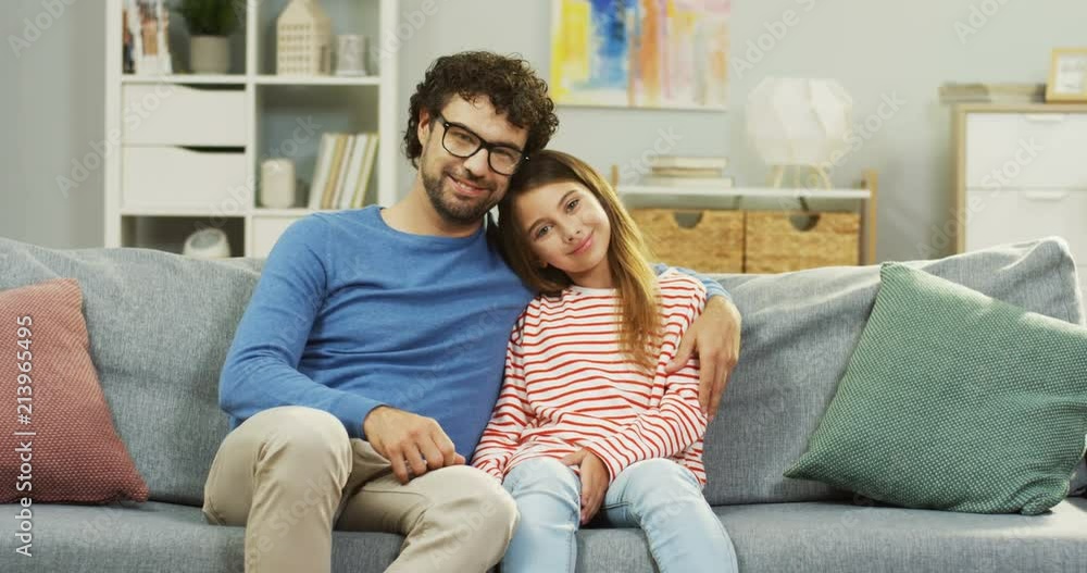 Portrait shot of the cute small girl and her good looking father posing to the camera while sitting on the sofa in the living room and hugging. Indoor. Portrait shot of the cute small girl and her good looking father posing to the camera while sitting on the sofa in the living room and hugging. Indoor.