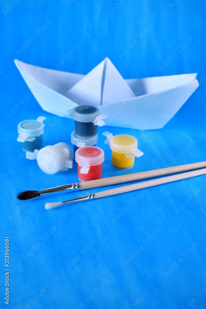 White paper ship, paints of different colours and brushes against the blue background White paper ship, paints of different colours and brushes against the blue background
