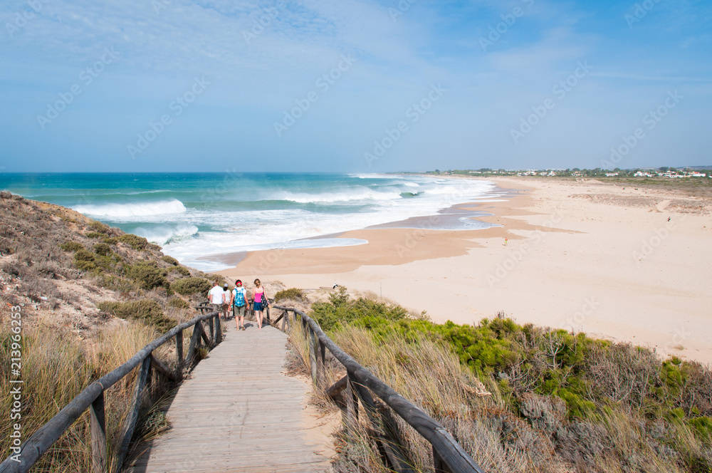 Group of tourists walking down boardwalk from Trafalgar lighthouse towards Zahora beach on sunny day in Andalusia, Spain. Summer vacation, travel destination, relax holidays concepts
