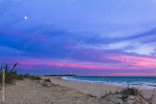 Colorful sunset with moon coming out and Trafalgar lighthouse on the background in empty Zahora beach  Cadiz  South Spain