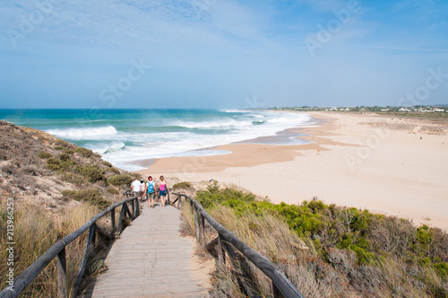 Group of tourists walking down boardwalk from Trafalgar lighthouse towards Zahora beach on sunny day in Andalusia  Spain. Summer vacation  travel destination  relax holidays concepts