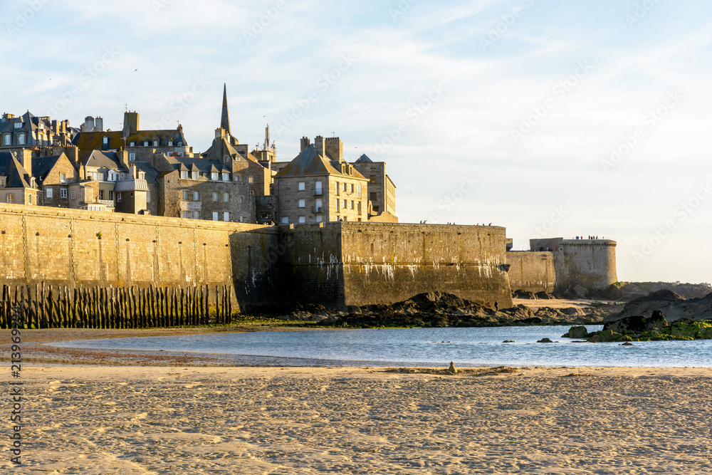 The walled city of Saint-Malo, France, at sunset with a spire sticking out from the buildings behind the wall, the Fort a la Reine, the Bidouane tower and a sand beach in the foreground.
