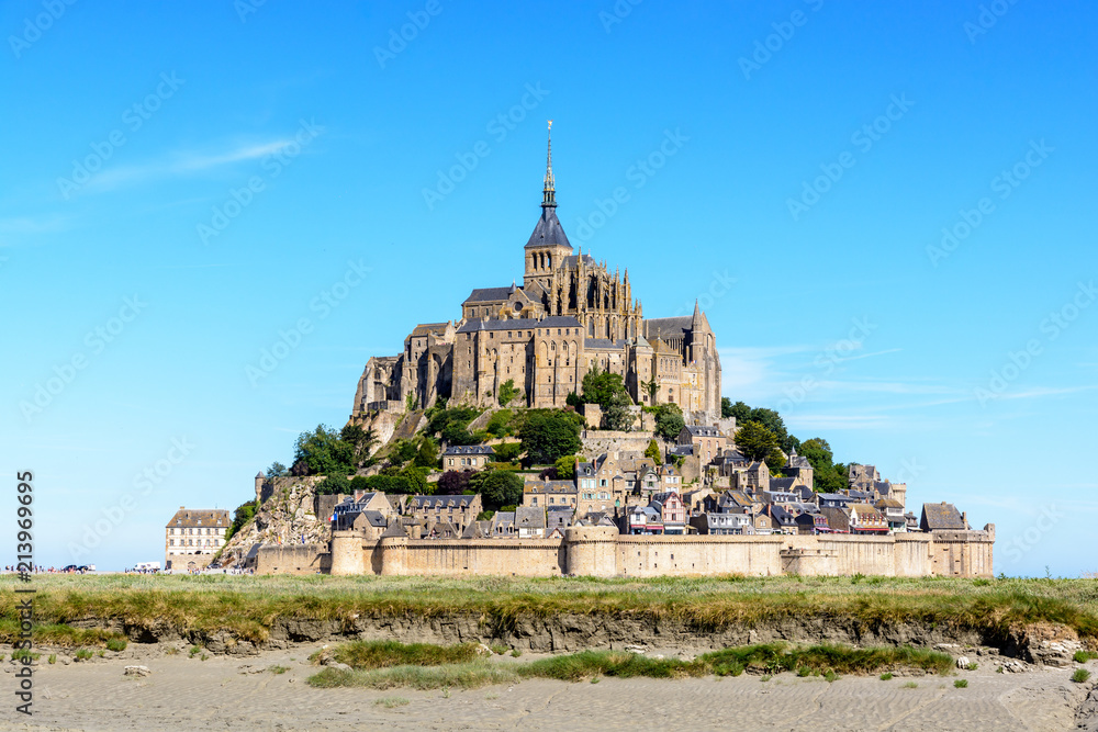 General view of the Mont Saint-Michel tidal island, located in France on the limit between Normandy and Brittany, from the bay at low tide under a summer blue sky with salt meadow in the foreground.