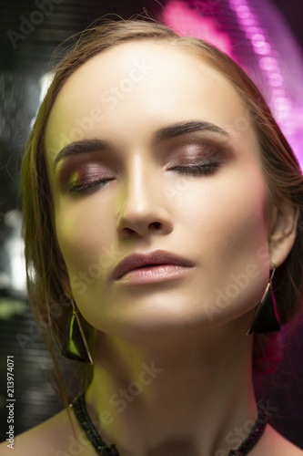 Beautiful girl with evening make-up wearing an elegant necklace and earrings of black opal on a background of a mirror disco ball  illuminated by green and pink lights