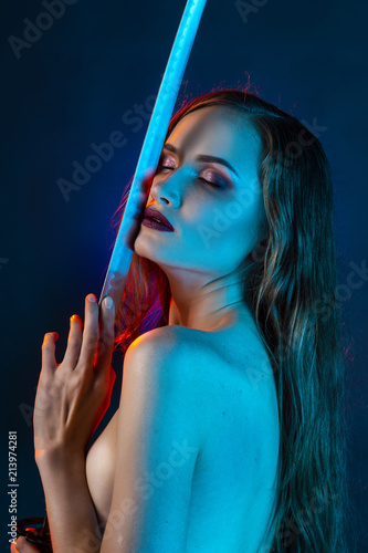 Beautiful girl wearing an elegant necklace and earrings of black opal holds Japanese katana sword in her hands on blue background, illuminated by red and blue lights