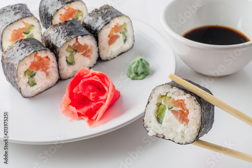 Sushi roll in chopsticks in soy sauce, closeup. Plate with roll japanes seafood sushi with ginger and wasabi on background.