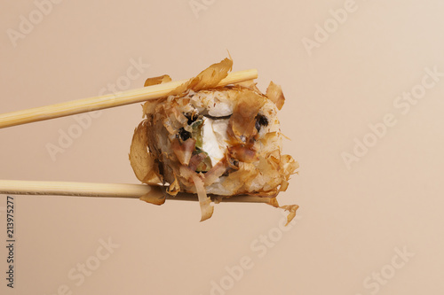 Fototapeta Naklejka Na Ścianę i Meble -  A Bonito roll with some cream cheese, a salmon and some smoked tuna shavings. close up of chopsticks taking portion of sushi roll on light background / eating sushi roll using chopsticks.