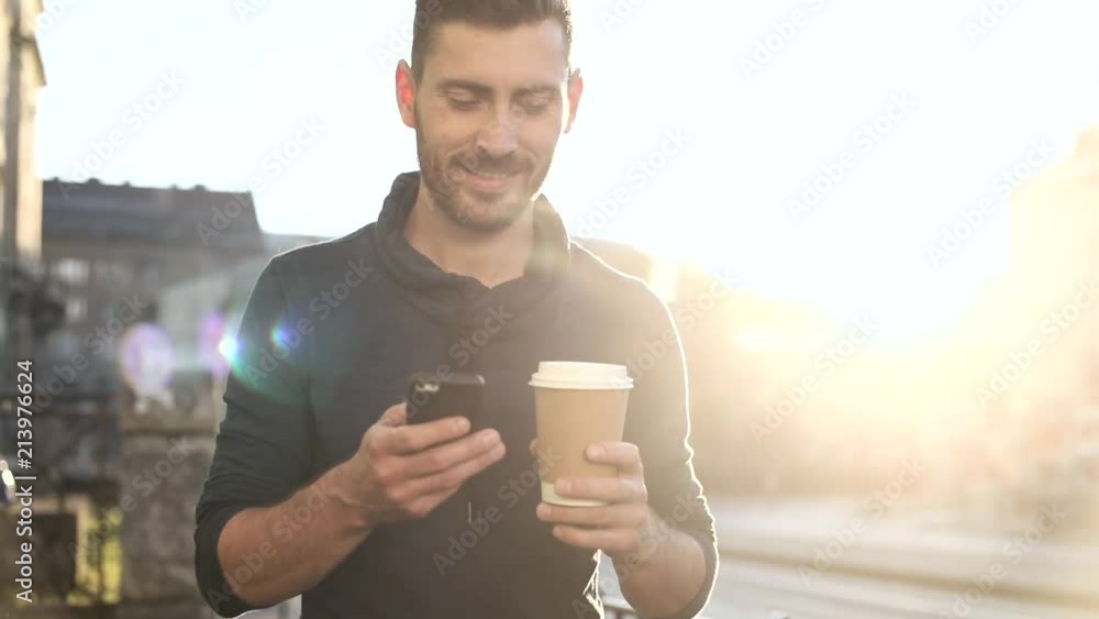 Happily Smiling Attractive Man Walking down the Street. Typing on his Smartphone with Interest. Enjoying the Evening Walk. Drinking Delicious Coffe. Crowded City on the Background. Happily Smiling Attractive Man Walking down the Street. Typing on his Smartphone with Interest. Enjoying the Evening Walk. Drinking Delicious Coffe. Crowded City on the Background.