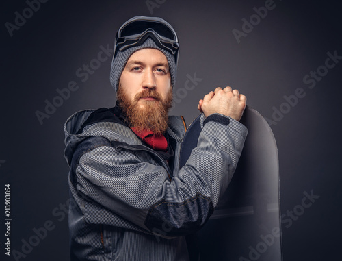 Brutal redhead snowboarder with a full beard in a winter hat and protective glasses dressed in a snowboarding coat posing with snowboard at a studio, looking away. Isolated on the gray background.