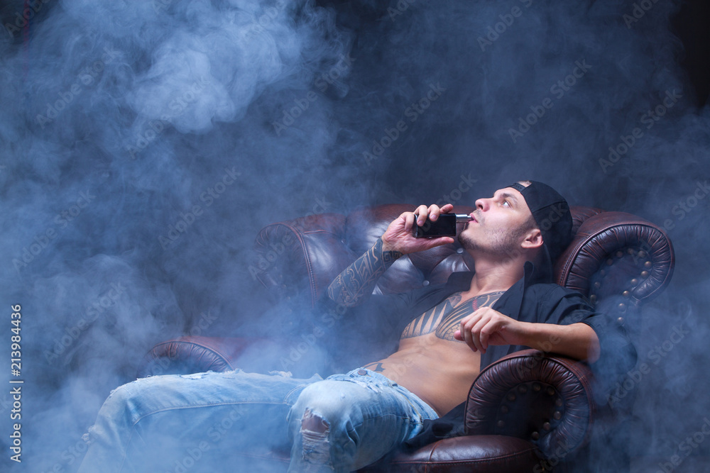 Vaper. The man with tattoos sits on a leather sofa smoke an electronic cigarette on the dark background.