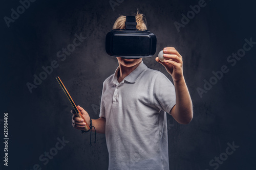 Blonde boy dressed in a white t-shirt playing ping-pong with a virtual reality glasses. Isolated on dark textured background.