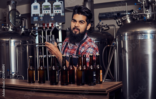 Stylish full bearded Indian man in a fleece shirt and apron holds a glass of beer, standing behind the counter in the brewery. © Fxquadro