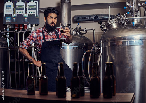 Stylish full bearded Indian man in a fleece shirt and apron holds a glass of beer, standing behind the counter in the brewery.