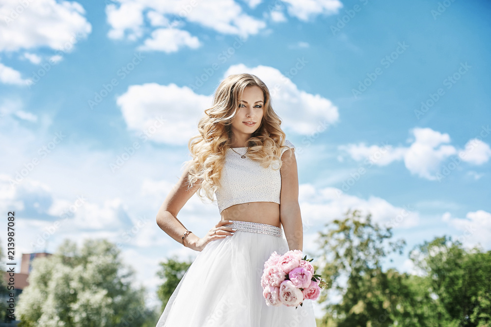 Beautiful and sensual blonde model girl with sexy body in stylish tulle skirt and in fashionable blouse, with bouquet of flowers in her hand posing outdoors with blue skies at background